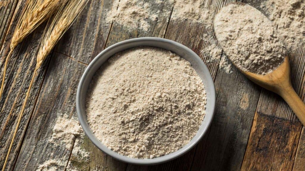 Whole wheat flour in a bowl and spoon beside it on table with 2 wheat plants beside it