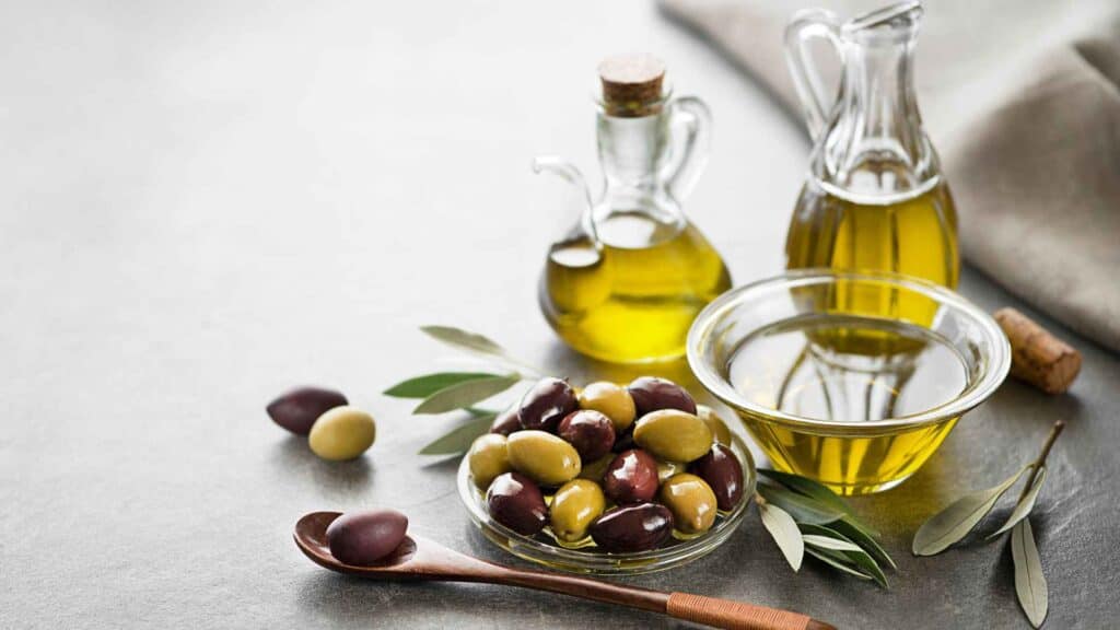 Olive oil in jug, bowl and olive seeds, leaves and wooden spoon on table